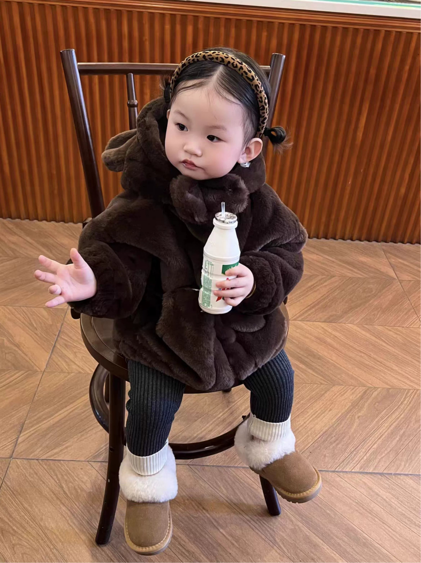 Child in a brown outfit sitting on a chair holding a bottle.