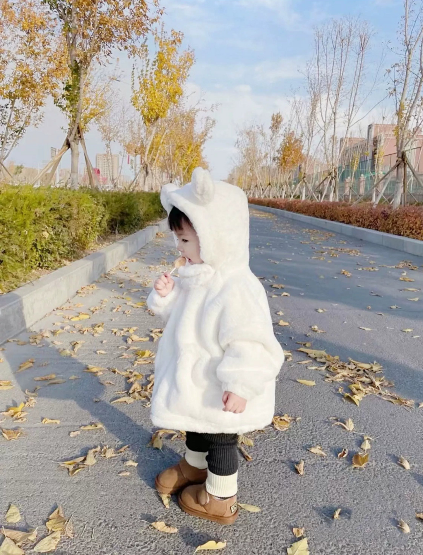 Child in a white hooded coat walking on a path with autumn leaves and trees in the background.