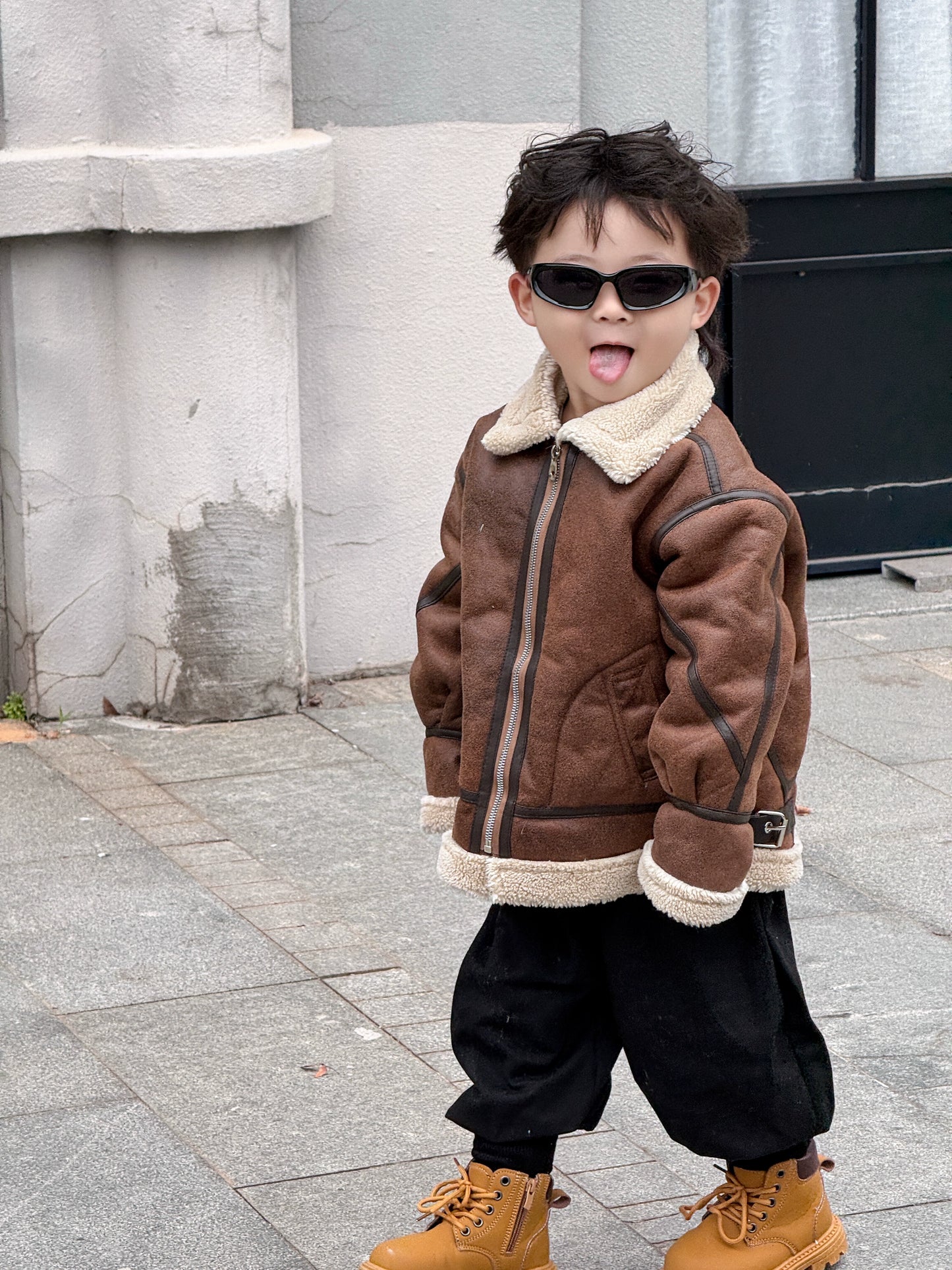 Child wearing a brown jacket with a fur-lined hood, black pants, and tan boots on a sidewalk.