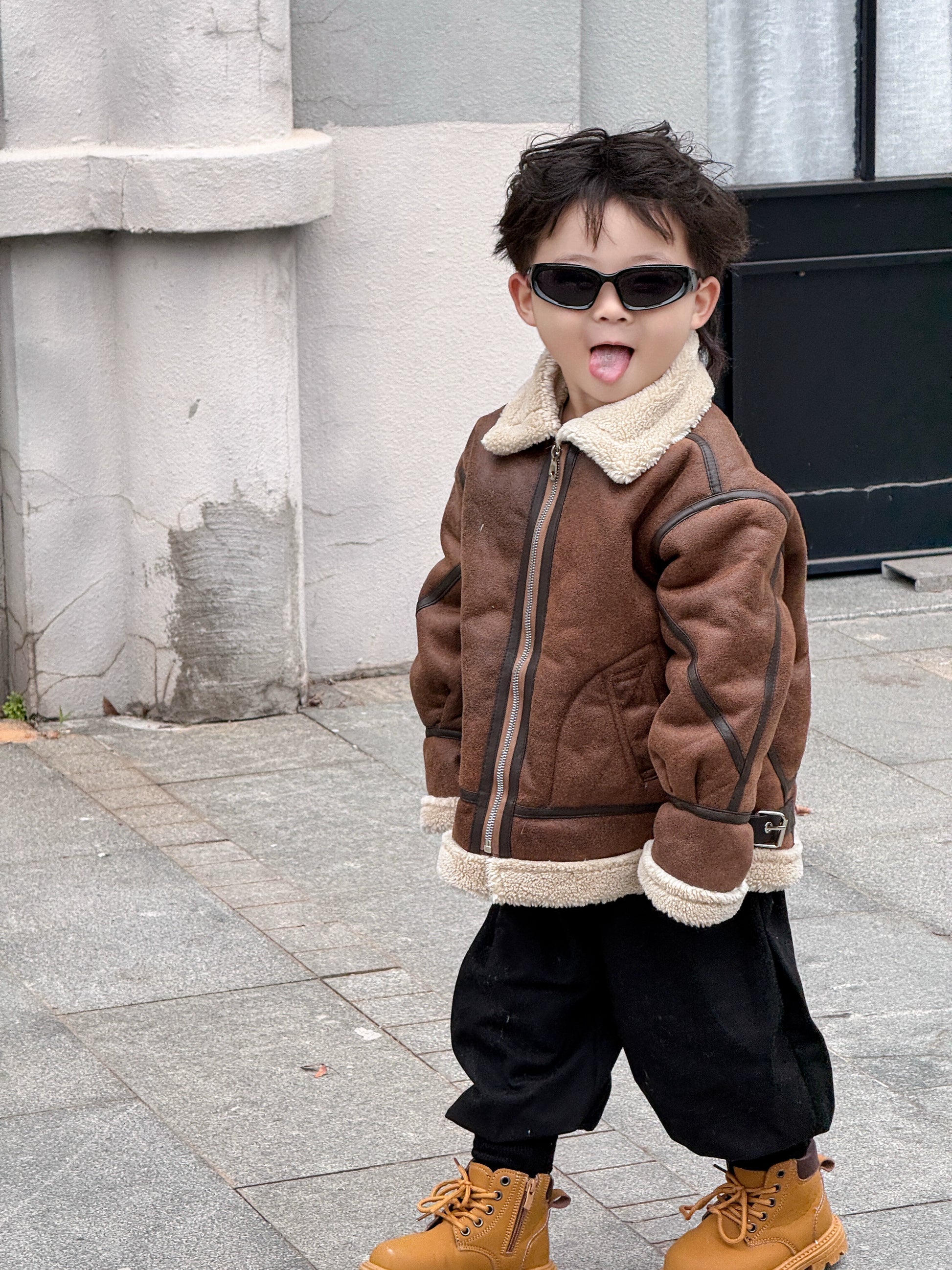 Child wearing a brown jacket with a fur-lined hood, black pants, and tan boots on a sidewalk.