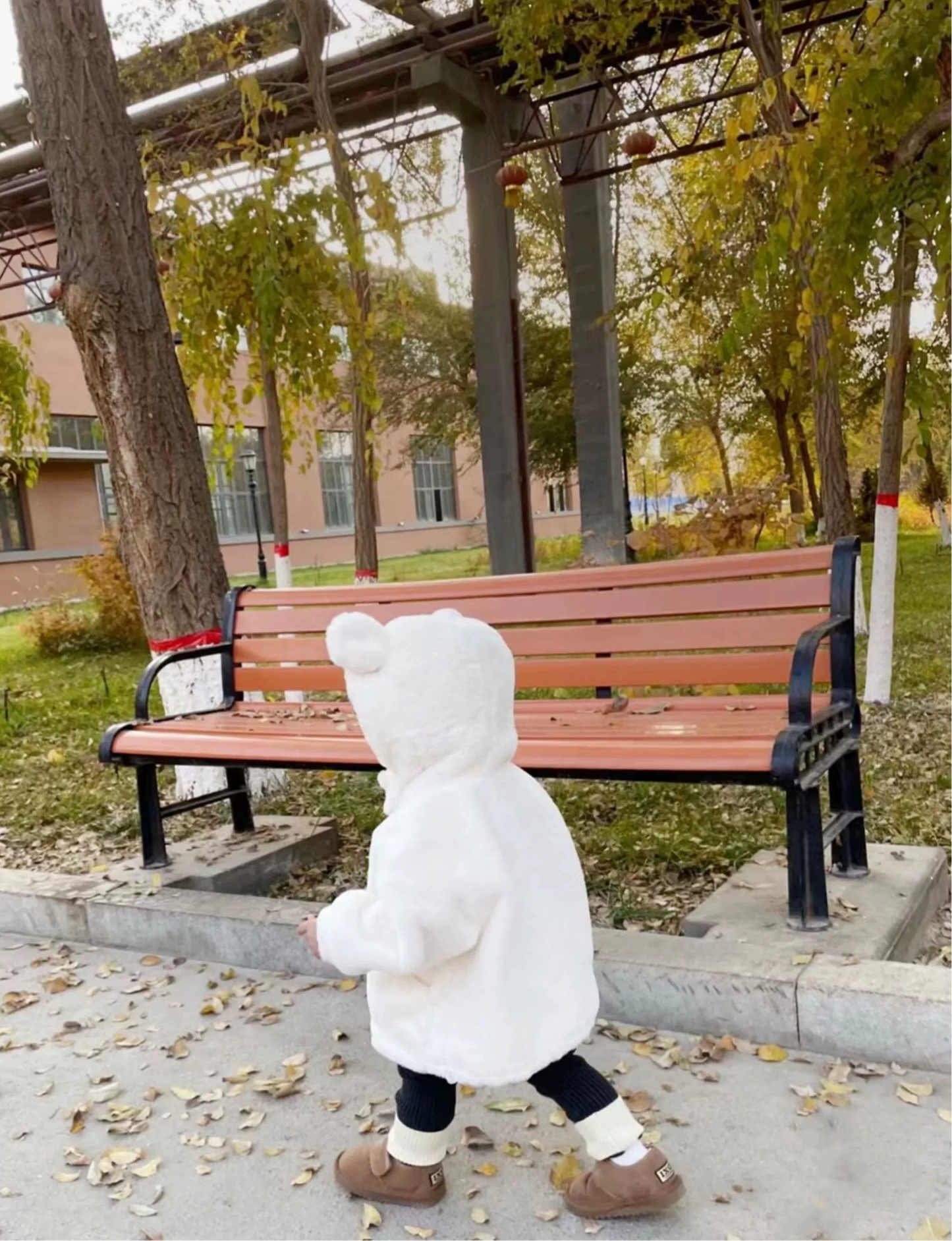 Child in a white coat standing on a sidewalk with a bench and trees in the background