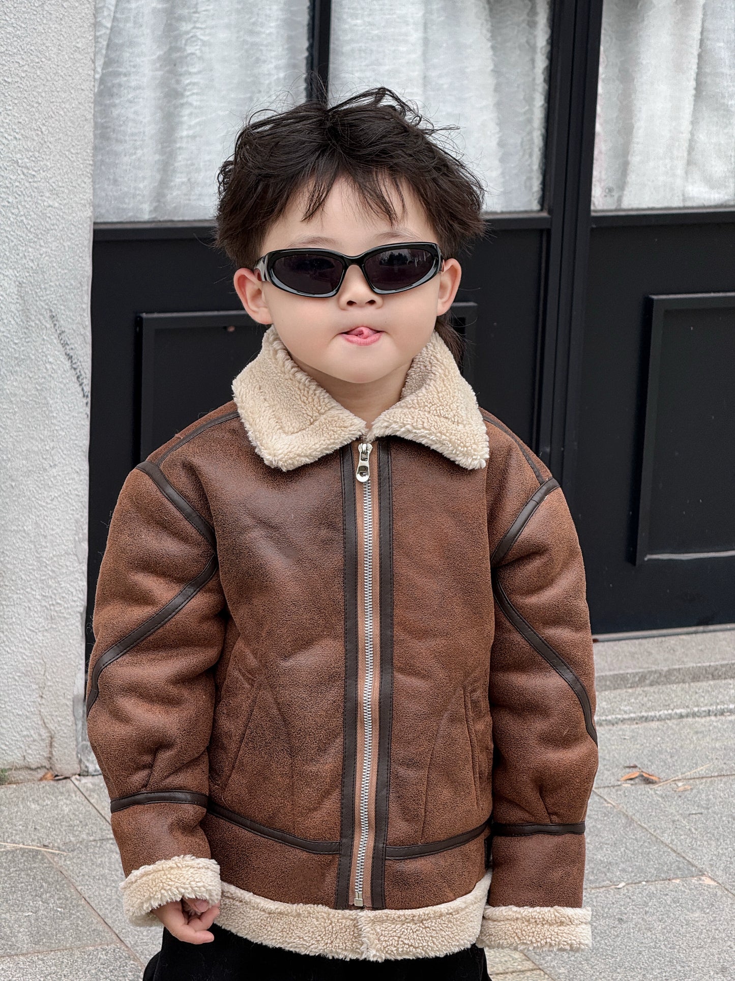 Child wearing a brown leather jacket with fur trim standing in front of a building entrance.
