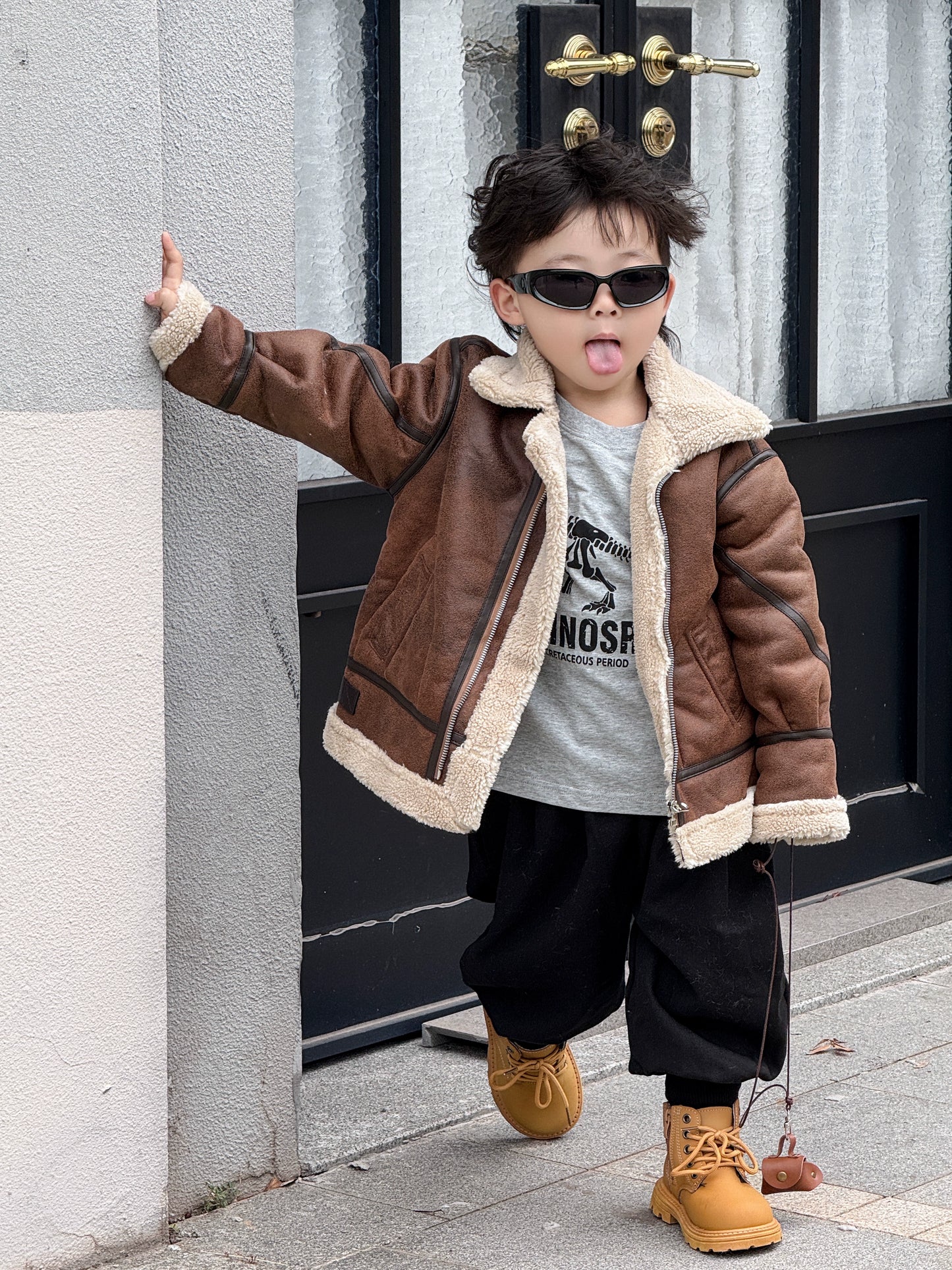 Child wearing a brown leather jacket with fur lining, sunglasses, and standing against a building.