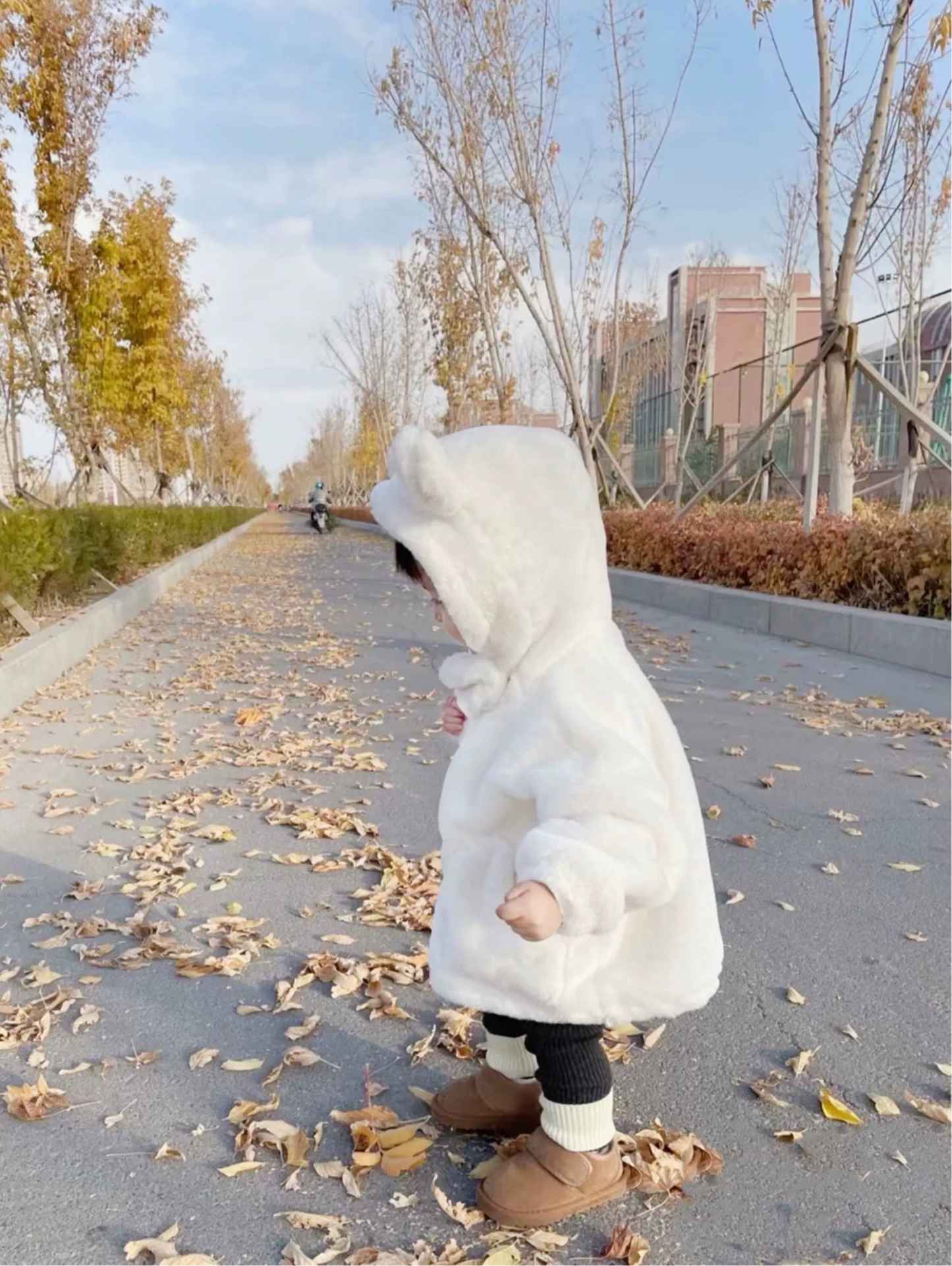 Child in a white hooded coat walking on a path with fallen leaves and trees in the background.