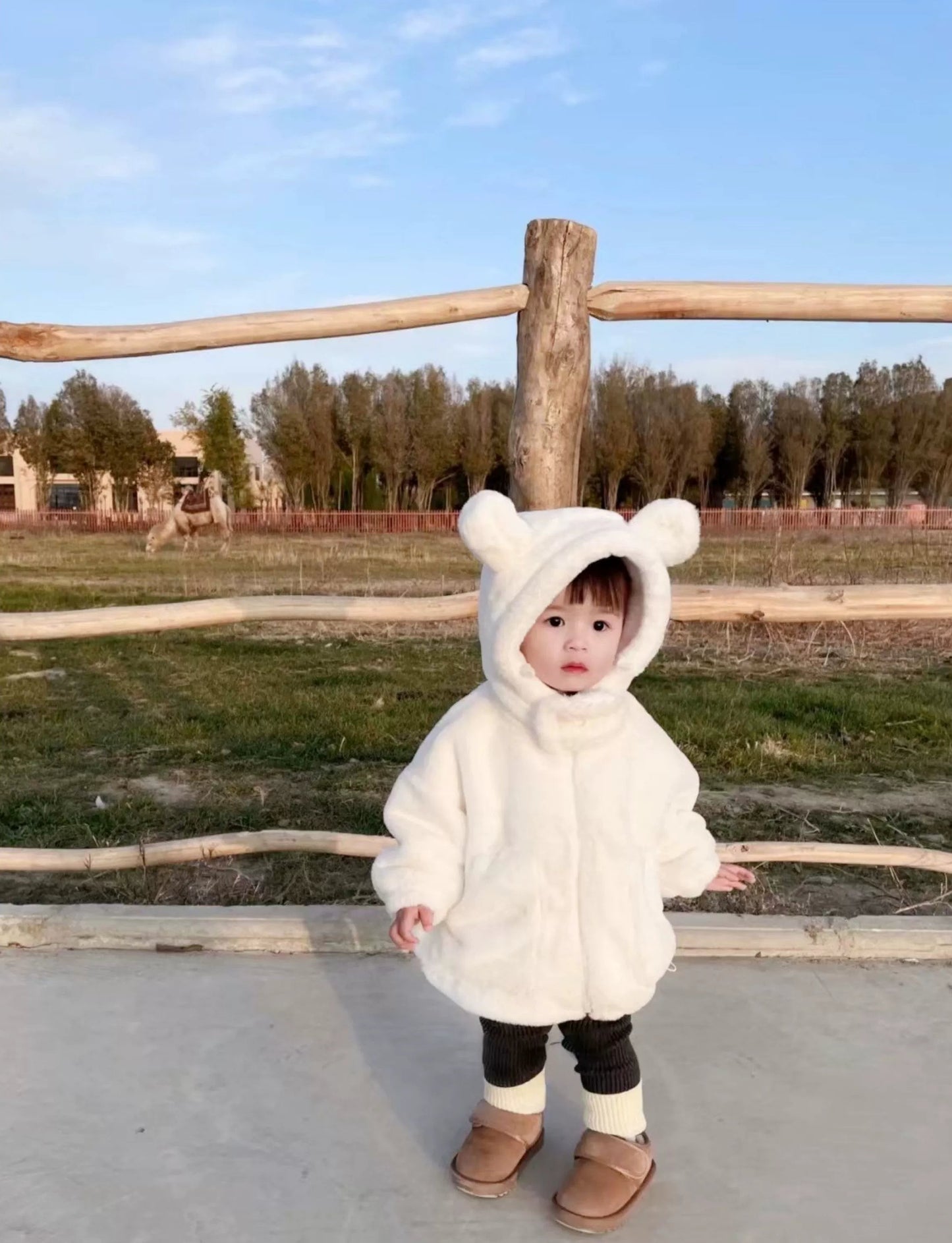 Child wearing a white bear-themed hoodie outdoors with a wooden fence and trees in the background.