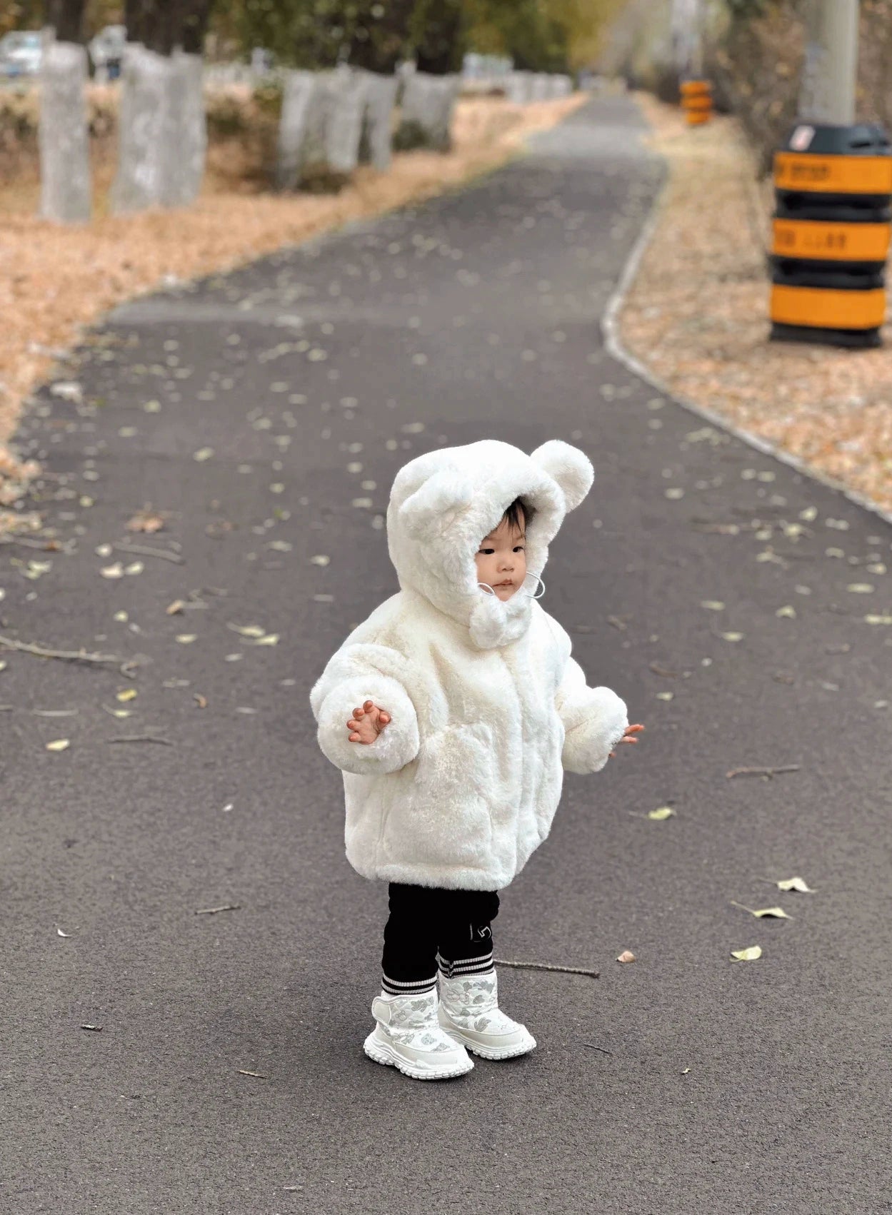 Child in a white fluffy coat standing on a paved path with trees and barriers in the background.