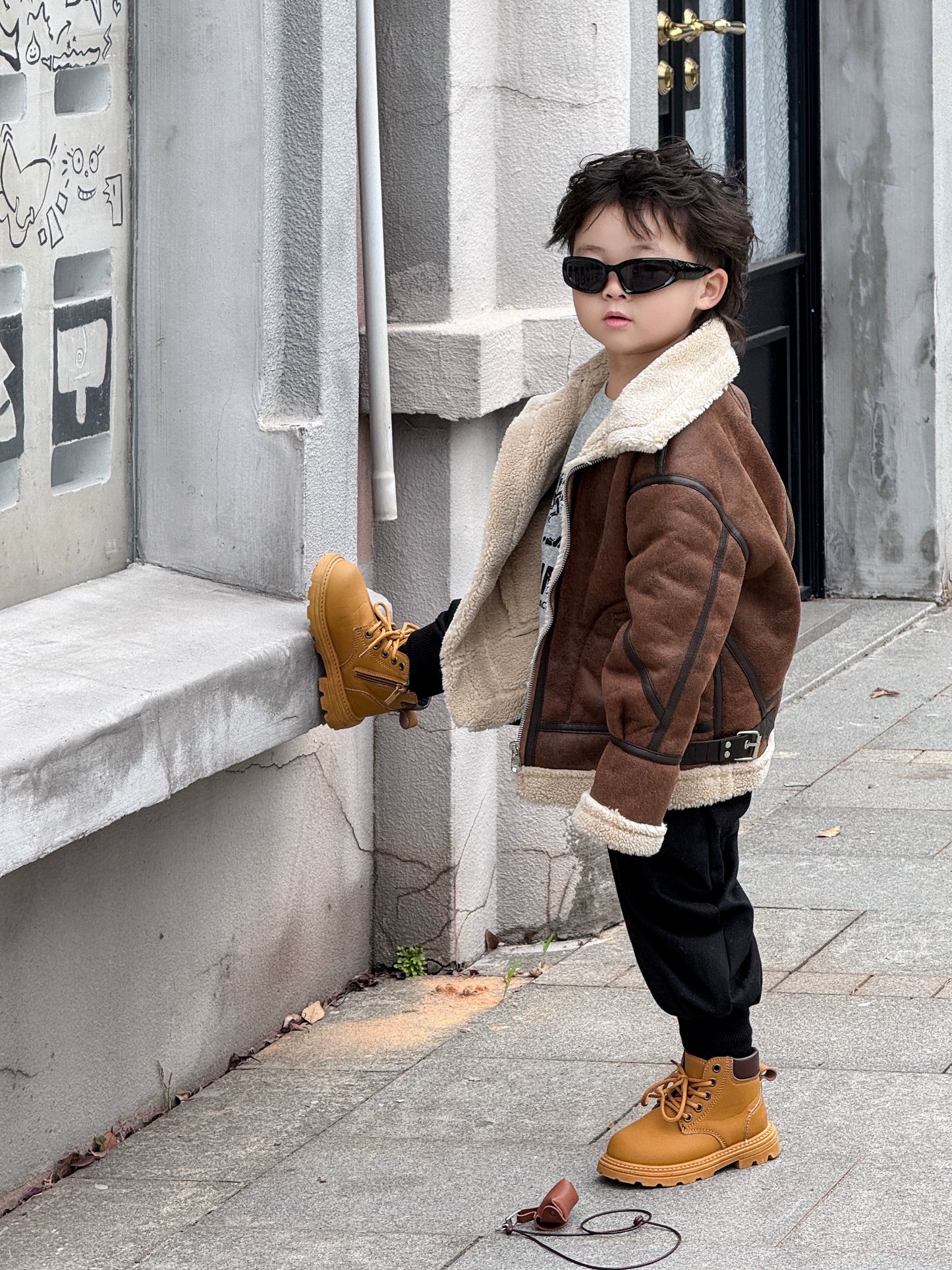 Child wearing a brown jacket and yellow boots standing on a sidewalk.
