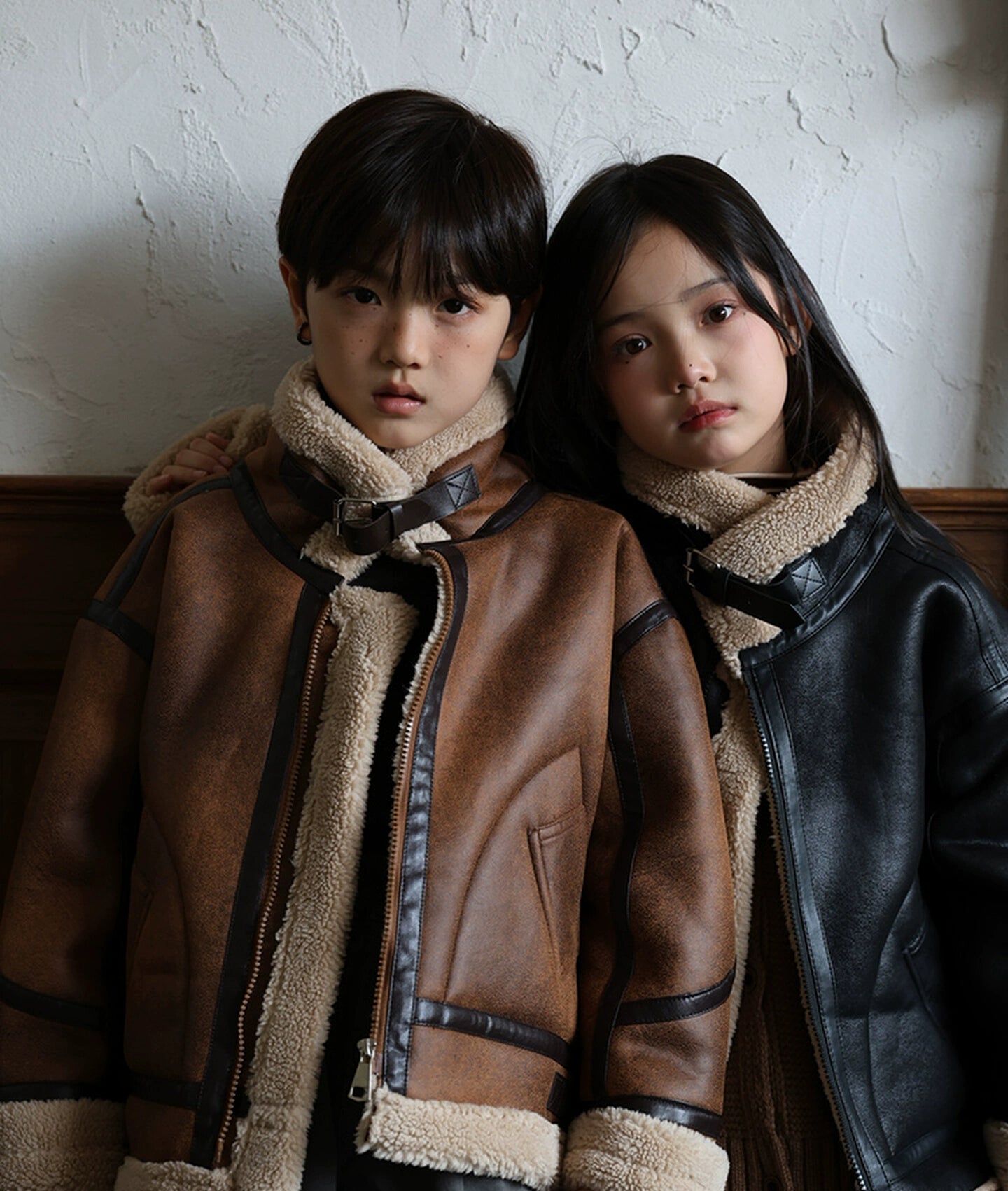 Two children wearing brown leather jackets with fur-lined hoods against a plain wall.