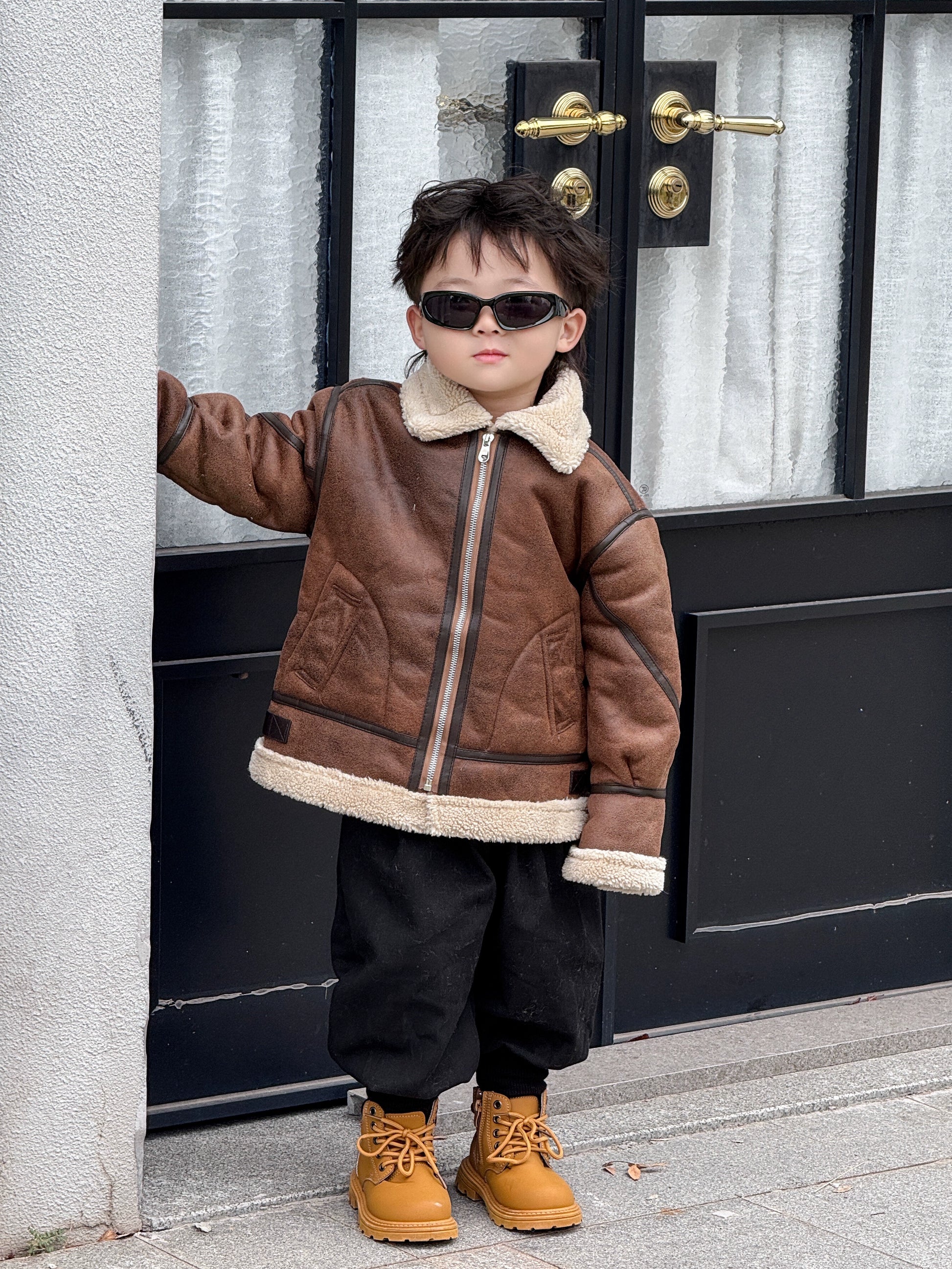 Child wearing a brown jacket with white fur trim, black pants, and tan boots standing in front of a door.