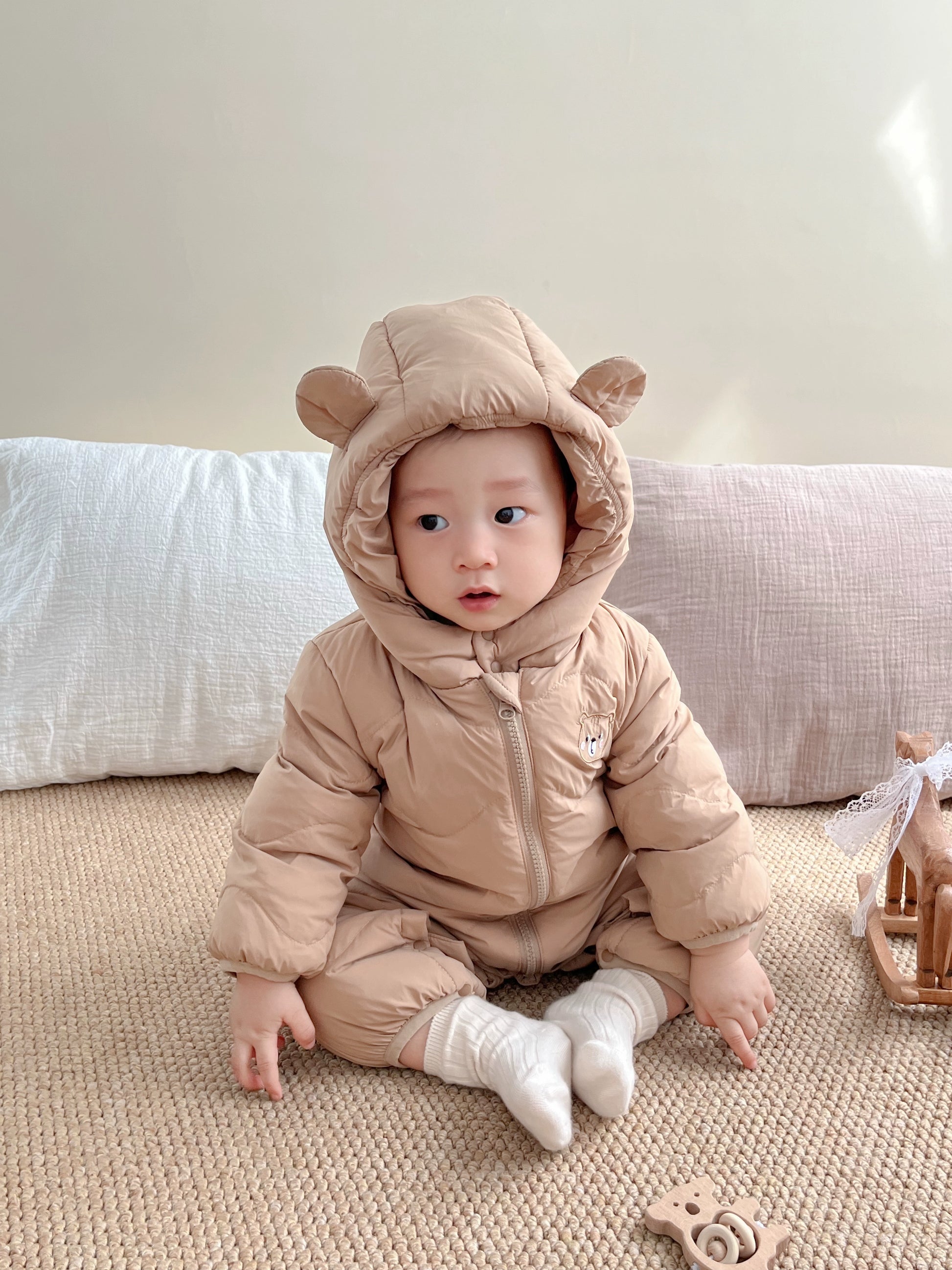 Baby wearing a beige bear-themed snowsuit sitting on a soft surface.