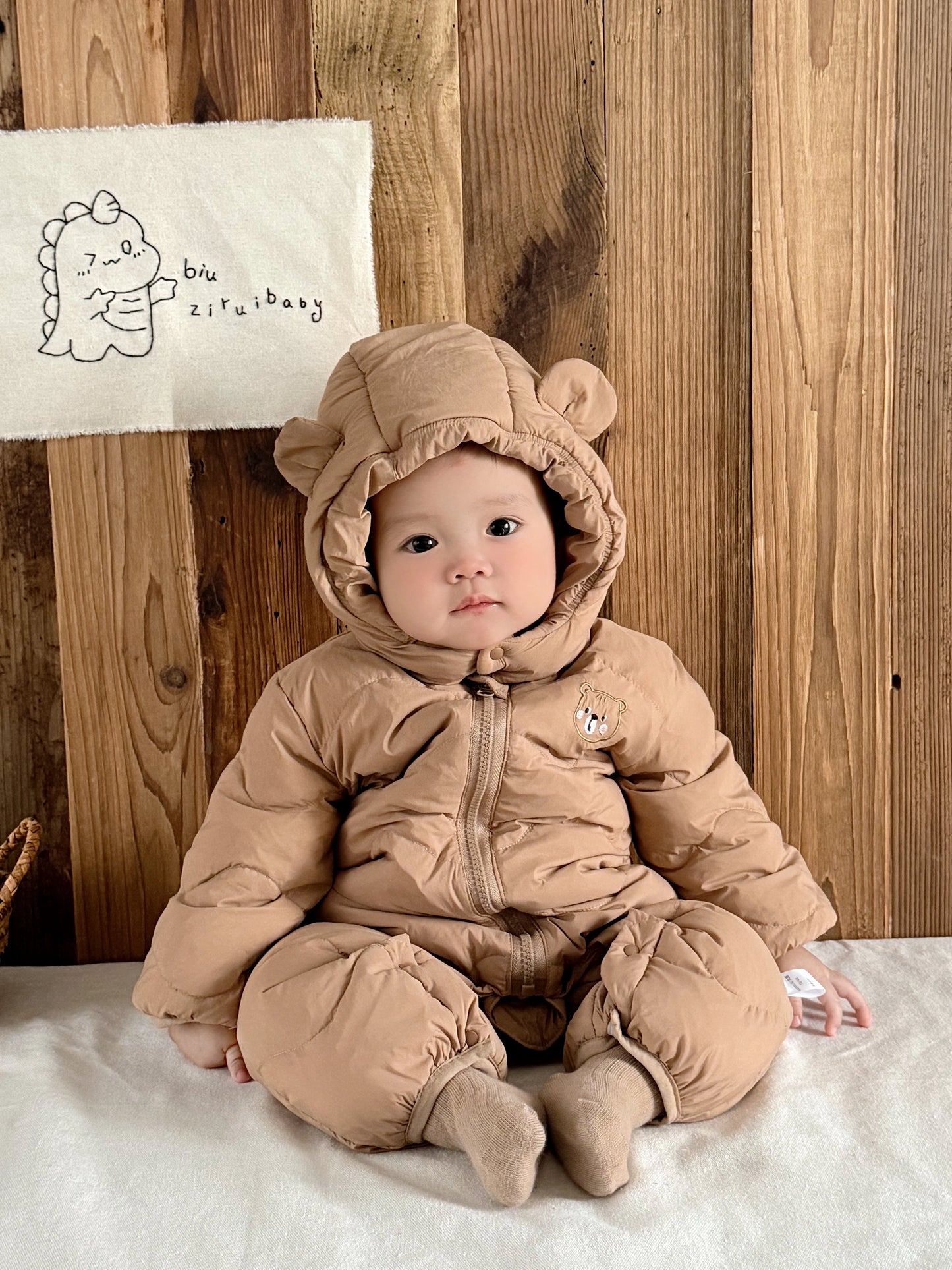 Baby in a beige snowsuit with bear ears sitting on a wooden floor.