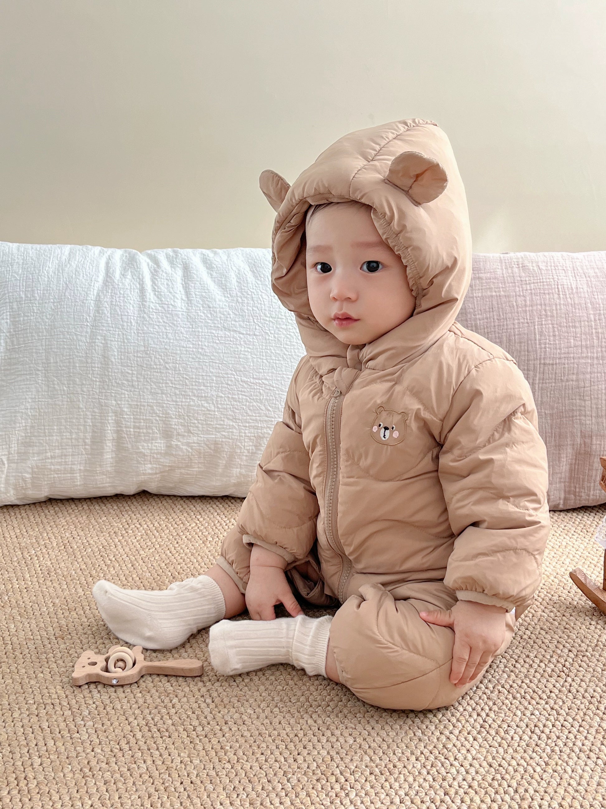 Baby in a beige snowsuit with bear ears sitting on a couch.
