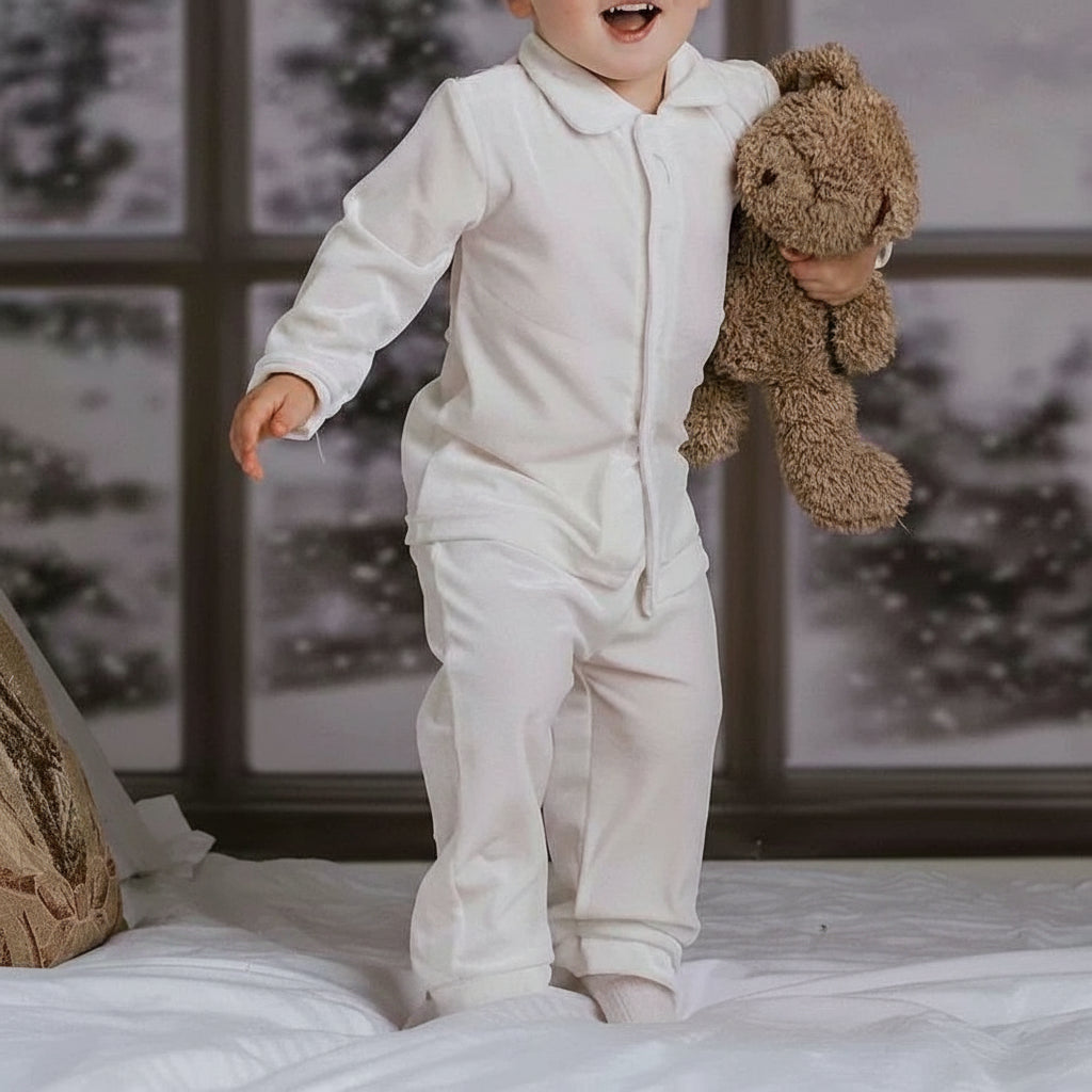 Child in a white onesie holding a brown teddy bear in a bedroom setting.