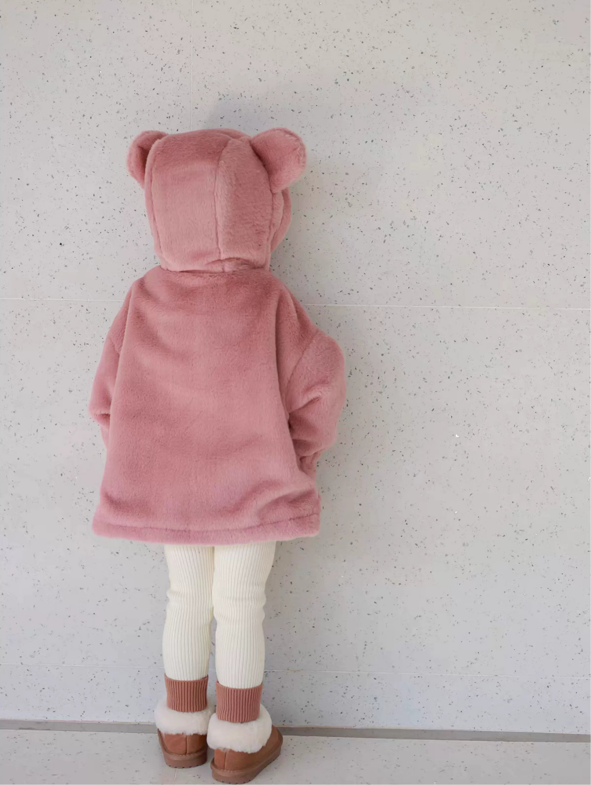 Child wearing a pink bear-themed coat against a light gray wall.