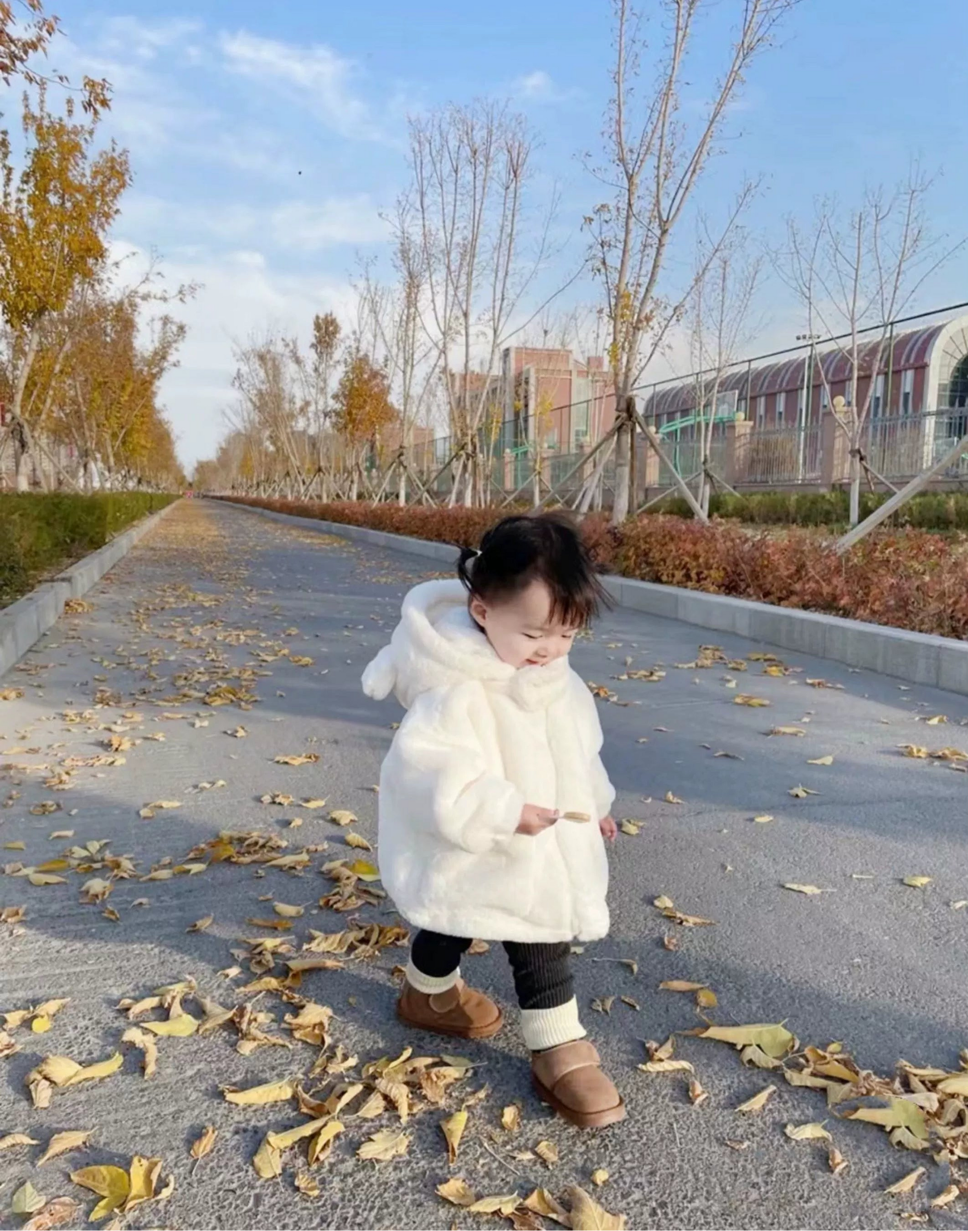 Child in a white coat standing on a path with autumn leaves and trees in the background