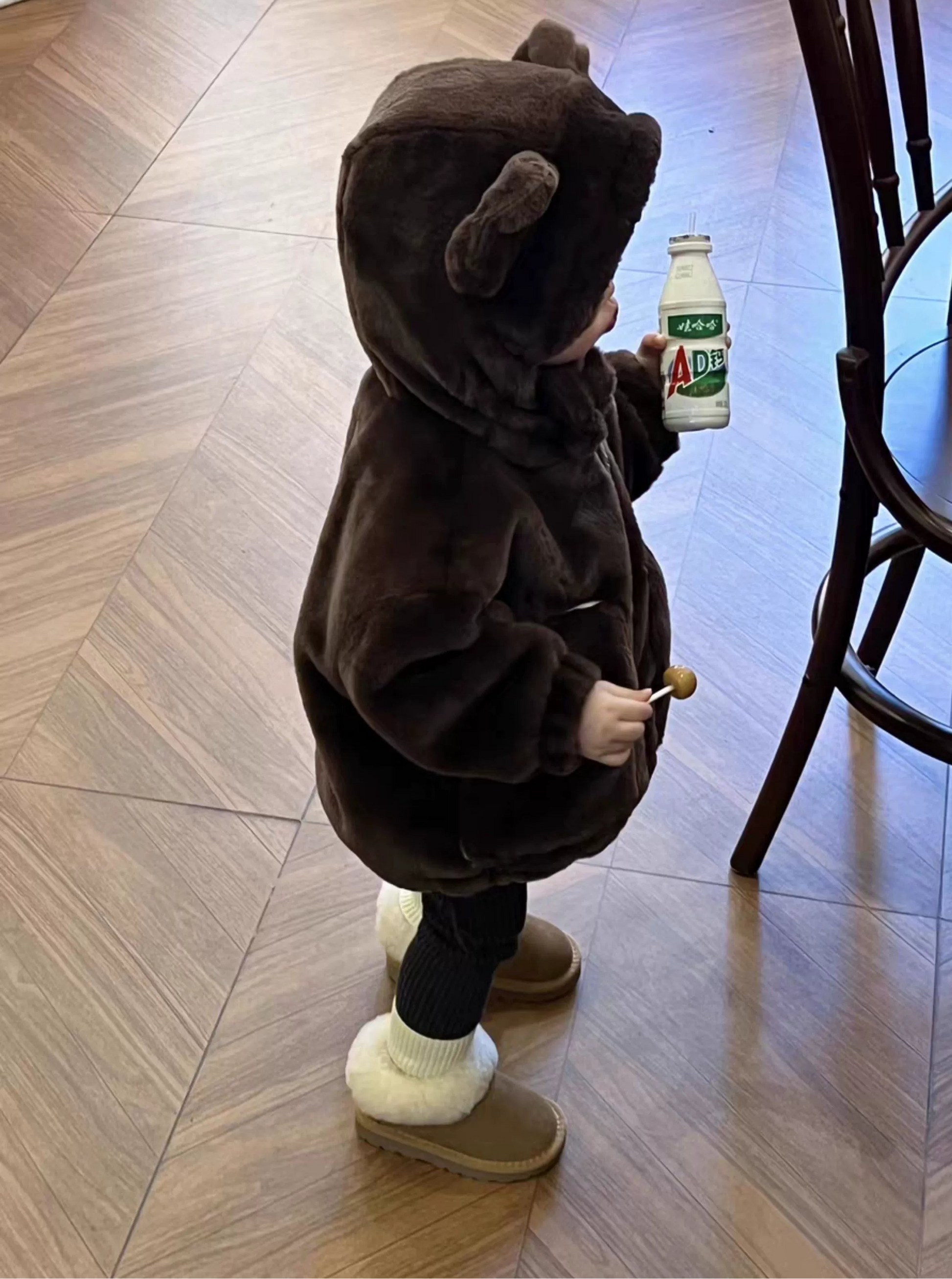 Child in a brown bear costume holding a bottle on a wooden floor.