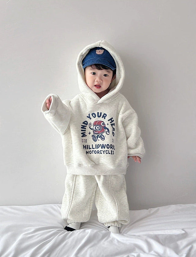 Child wearing a white hoodie and pants with a graphic design, standing on a bed against a plain wall.