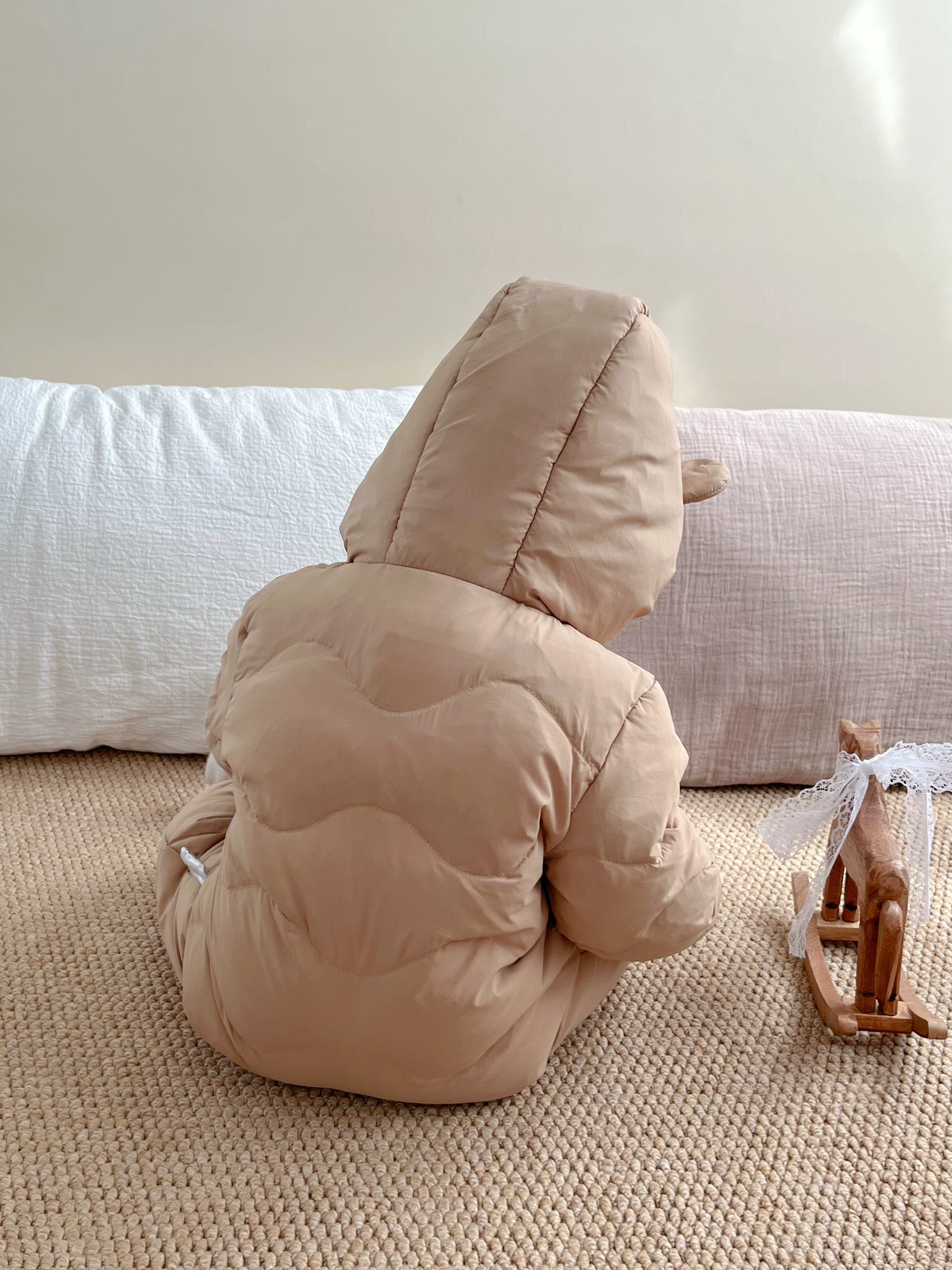 Child in a beige hooded outfit sitting on a carpeted floor with a white wall in the background.