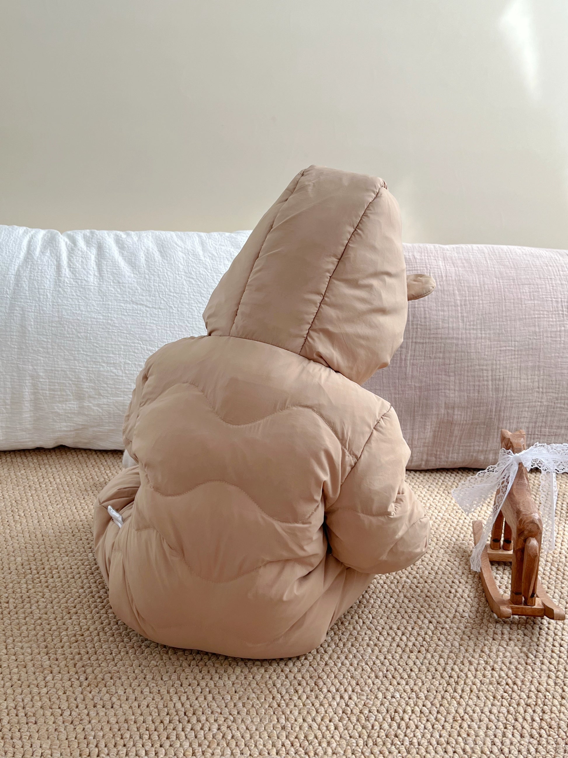 Child in a beige hooded outfit sitting on a carpeted floor with a white wall in the background.