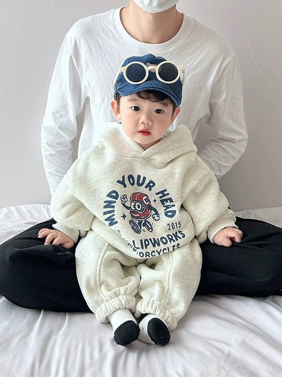 Child wearing a white hoodie with text and graphics, sitting on a bed with an adult behind.