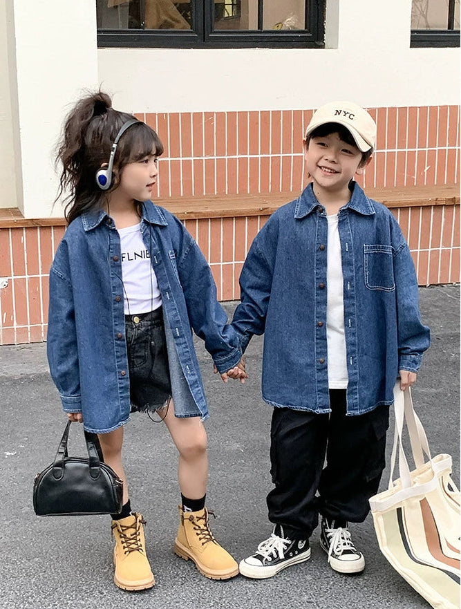 Two children holding hands, both wearing denim jackets and smiling outdoors.